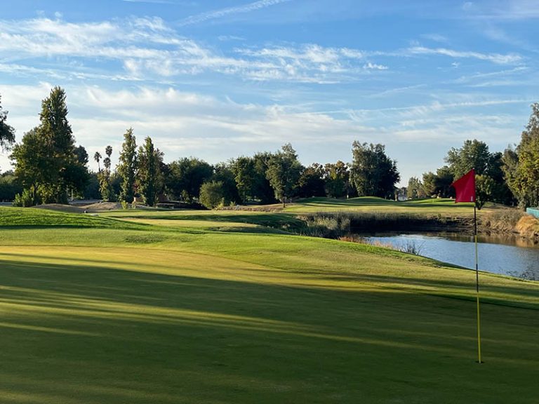 Red flag on a Lemoore Golf Course green beside water with fairway and trees in the background.